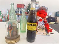 Group shot showing Coca-Cola bottle, NuGrape bottle, John Wayne dome figurine, boxed plush frog