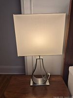 Lamp front view showing white square shade illuminated and brushed nickel open ribbon base