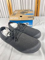 Side view of both black hemp canvas shoes with laces, stuffing and damaged retail box in background