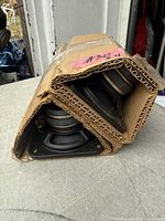 Pair of square speaker drivers visible inside folded cardboard, showing magnets and mounting frames