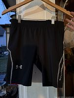 Front view of black Under Armour youth compression shorts hanging, logo on left leg, vertical branding on right leg visible.