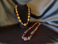 Both necklaces displayed: amber-brown graduated bead strand on bust, red-brown mixed bead strand laid flat
