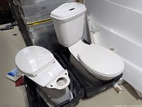 Two white toilet bowls in plastic bins; right bowl has tank, left missing tank