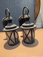 Two black metal and glass lantern wall sconces with curved arms and mounting backplates