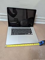 MacBook Pro open showing keyboard and screen with tape measure indicating width ~14.5 inches