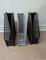 Front view of three metal mesh magazine holders (two black, one silver) standing upright.