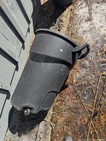 Side view of Rubbermaid black wheeled garbage can leaning against wall, showing wheels and handle