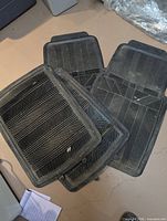 Stack of four black rubber car floor mats displaying tread patterns and size difference