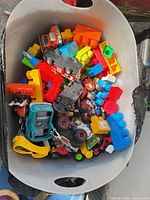 Bin containing assorted plastic toy cars, trucks, building blocks, and hand tools