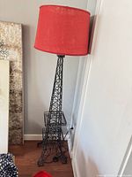 Full view of black metal Eiffel Tower floor lamp with red drum shade against wall