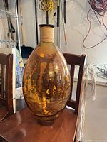 Full view of the large amber hand-blown glass vase on chair, showing color, texture, and rope-wrapped neck.
