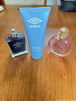 Three Umbro personal care items on wooden surface: navy men’s cologne bottle without cap, blue shower gel tube, pink women’s perfume bottle with cap