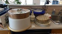 Countertop view with cake carrier, wood bowl, Pampered Chef chopper, boxed appliance, glass pie plate and other kitchen items