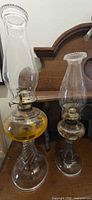 Both clear glass oil lamps side by side showing bases, reservoirs, chimneys and brass fittings