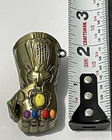 Front view of metal Infinity Gauntlet charm beside ruler