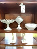 Three milk glass pieces on upper shelf: pedestal compote, lidded candy dish, scalloped bowl; two twisted column sculptures visible on lower shelf