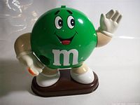 Front view of green M&M candy dispenser showing character face, waving arm, brown base