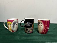Front view of three ceramic mugs: two Laura Secord portrait mugs and one black hummingbird mug