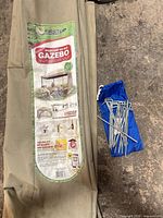 Folded tan canopy with FirstUp label beside blue pouch holding metal stakes