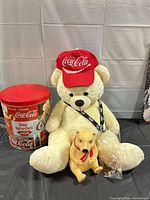 Lot overview: plush teddy bear wearing Coca-Cola hat, tin, money bank, lanyard