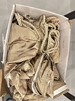 Box containing folded gold silk-like drapery panels and braided rope tiebacks