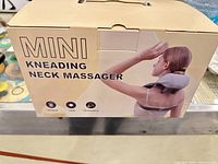 Retail box labeled Mini Kneading Neck Massager showing gray device on model; icons for wireless, heat, 3D kneading visible