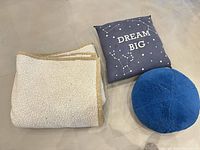 All three items laid flat: folded sherpa throw, blue pillow with DREAM BIG, round pouf