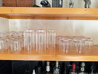 Row of inverted clear plastic stemmed wine glasses and tumblers on wooden shelf