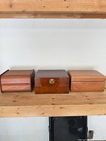 Three wood jewelry boxes on shelf: square burl box center, two rectangular wood boxes left and right