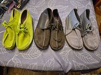 Three pairs of Clarks suede lace-up shoes in lime green, brown and grey displayed side by side on table