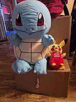 Large Squirtle plush seated next to smaller Pikachu on cardboard box