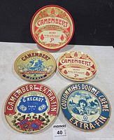 Five round items displayed; four ceramic plates with distinct cheese-label designs plus presentation box lid