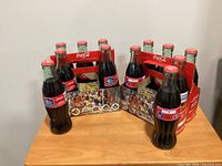 Two 6-pack carriers with twelve sealed commemorative Coca-Cola bottles displayed on table