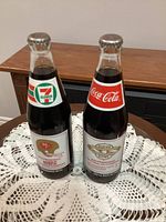 Front view of both sealed commemorative Coca-Cola bottles showing labels and silver caps.