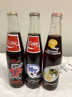 Front view of three sealed 10 oz Coca-Cola NCAA bottles showing team logos