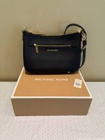 Front view of black leather Michael Kors crossbody bag on top of branded box with tag