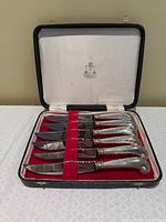 Open case showing six knives with silver-plated handles in red interior