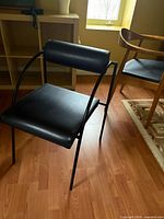 Front angle view of one chair showing black vinyl seat, cylindrical backrest, curved metal arms