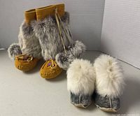 Front view showing both pairs: tall tan moccasins with grey fur and pom-poms; blue suede baby slippers with white fur cuffs