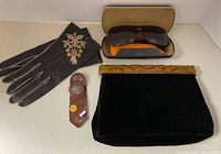 Group shot showing clutch, brooch on gloves, sunglasses with case, leather gloves, enamel pins