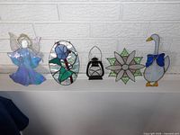 Five stained glass pieces on ledge: angel, blue rose oval, lantern, eight-point star, goose