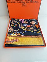 Scarf folded in orange Hermès box showing black ground, multicolour design, yellow border