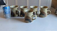 Five matching stoneware coffee mugs beside soda can for scale