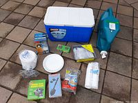 All items laid out: cooler, water container, boxed lantern, folding stove, folding shovel, Off! candles, disposable bowls, plate, cutlery set, cups, camp soap