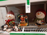 Shelf view showing boxed Coca-Cola Santa bank, snowman string lights, two plush snowmen
