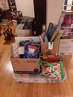 Lot overview showing rolls of wrapping paper, gift bags in cloth totes, cardboard box of kraft bags, all atop clear plastic bin