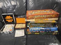 Stack of five board game boxes with two domino sets, including aluminum Mexican Train case, displayed on sofa