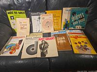 Group shot of approximately 15 music books and sheet music laid on a sofa, titles visible