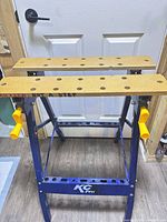 Front view of blue KC Pro folding workbench showing MDF jaws, bench dog holes and yellow crank handles