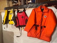 Overall view of three flotation devices hanging: Fluid yellow/black vest, WindRiver red/black vest, orange buoyancy coat